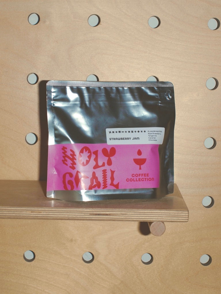 Front of an 8 oz bag of Strawberry Jam Colombia Santa Mónica from WeBe Coffee Roasters, silver pouch with pink Holy Grail Coffee Collection label, on a wooden pegboard shelf.