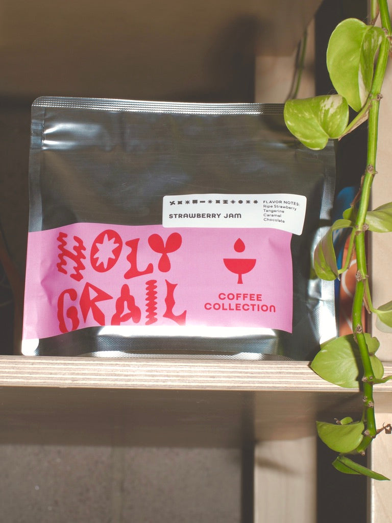 
                  
                    Front of the WeBe Coffee Roasters Strawberry Jam Colombia Santa Mónica bag with pink Holy Grail label and flavor notes, on a wood shelf beside a trailing pothos plant.
                  
                