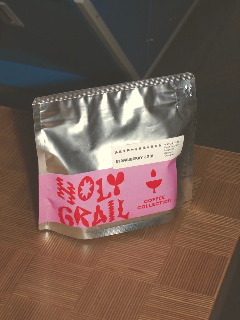 
                  
                    Silver bag of WeBe Coffee Roasters Strawberry Jam Colombia Santa Mónica with pink Holy Grail label, angled on a wood-tile tabletop.
                  
                