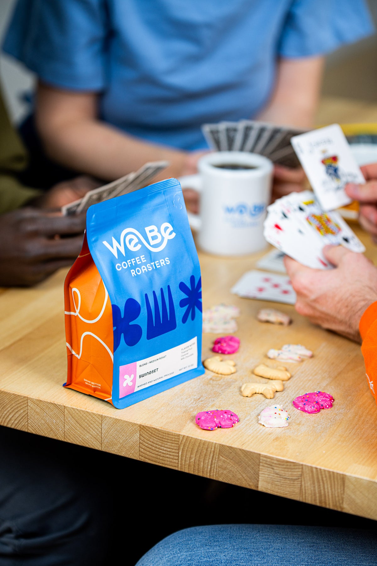 Friends playing cards around a bag of WeBe Swingset Blend and mugs of coffee