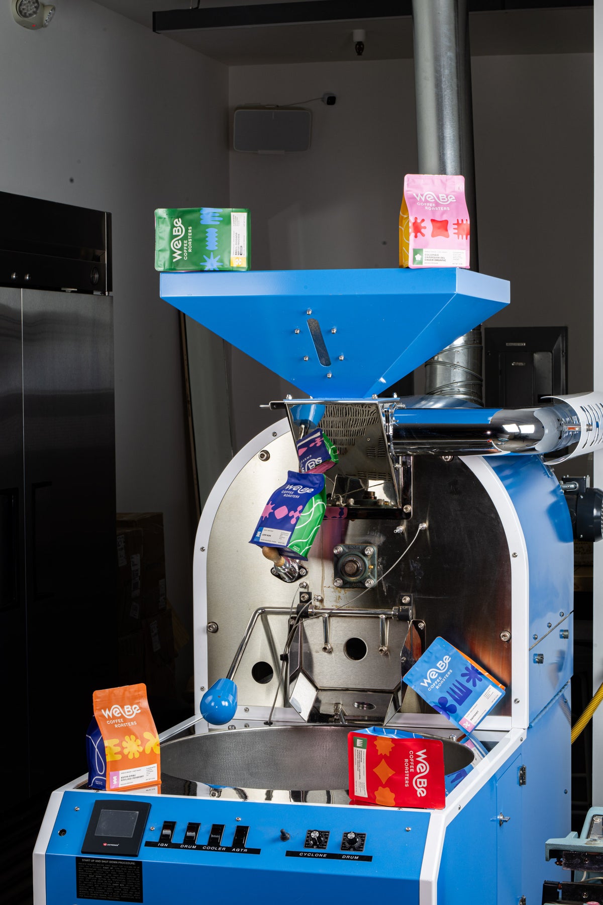 WeBe's blue coffee roaster with colorful bags of WeBe coffee